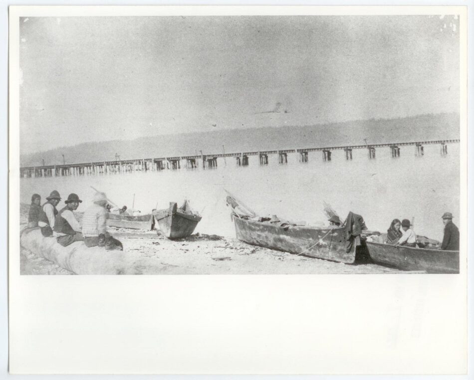 Old black and white photo of Puyallup Tribe sitting on beach with canoes